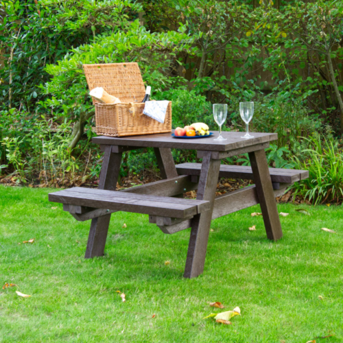 Small brown picnic table on grass with a picnic hamper, food and wine glasses set on it