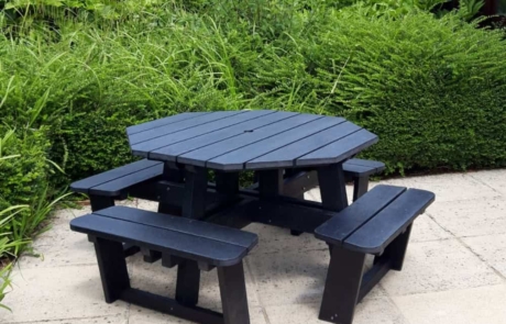 Octagonal-Picnic-Table-Black- Octagonal picnic table with seats on a patio with a hedge behind it.