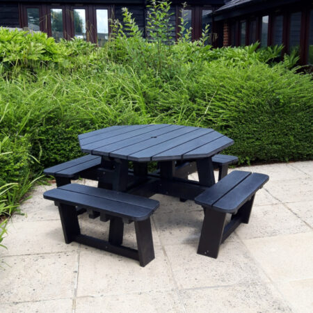 Octagonal Picnic Table in Black Octagonal picnic table with seats on a patio with a hedge behind it.