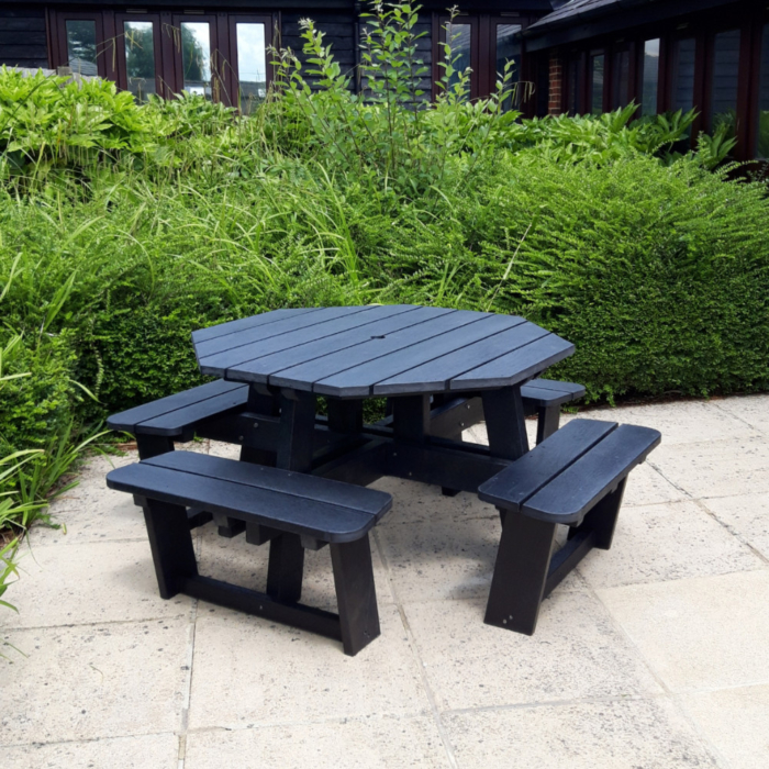 Octagonal picnic table with seats on a patio with a hedge behind it.