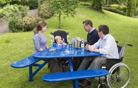 Blue steel triangular mobility picnic table in an outdoor setting with two people on seats, one child in a buggy and a a man in a wheelchair seated at it.