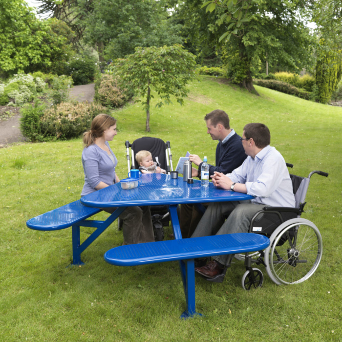 Blue steel triangular mobility picnic table in an outdoor setting with two people on seats, one child in a buggy and a a man in a wheelchair seated at it.